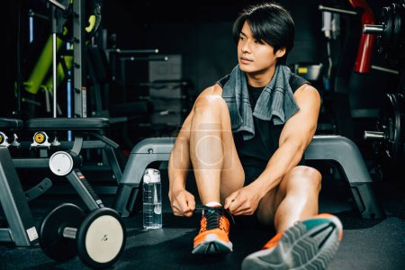 Photo for A fit and healthy adult man tying his shoelaces in his home gym. emphasizes his dedication to fitness and healthy living and includes a water bottle to remind viewers to stay hydrated during exercise - Royalty Free Image