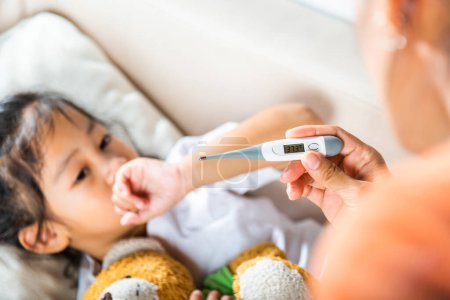 Photo for Sick kid. Mother parent checking temperature of her sick daughter with digital thermometer in mouth, child laying in bed taking measuring her temperature for fever and illness, healthcare - Royalty Free Image