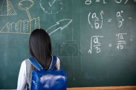 Photo for Education. Back view of school girl on science lesson in classroom write an answer on blackboard, primary child is standing in front of class in school writing chalkboard, Back to school concept - Royalty Free Image