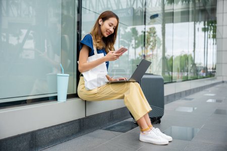 Photo for A woman with a beautiful smile working remotely while sitting on a bench at a train station. Her laptop and luggage are by her side as she balances work and travel. life of a modern entrepreneur. - Royalty Free Image