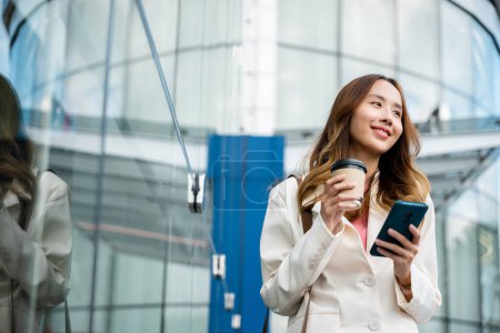 Téléchargez les photos : Young business woman smiling holding mobile phone with coffee take away going to work early in morning, Asian businesswoman with smartphone and cup coffee standing against street building near office - en image libre de droit