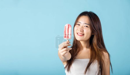Photo for Sweet tasty frozen dessert on summer time. Portrait of happy Asian young beautiful woman smiling and holding delicious ice cream wood stick mixed fruit flavor, studio shot isolated on blue background - Royalty Free Image