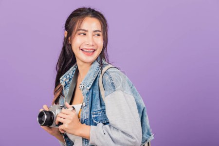 Téléchargez les photos : Attrayant portrait heureux asiatique belle jeune femme souriant excité usure denims et sac tenant vintage appareil photo, voyageur femme photographe, studio tourné isolé sur fond violet - en image libre de droit