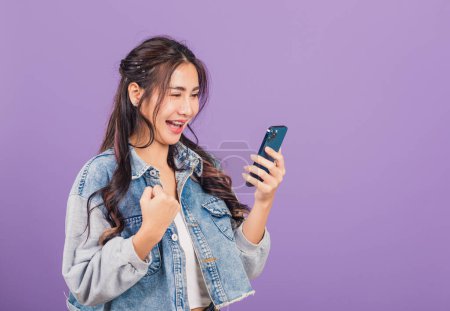 Téléchargez les photos : Portrait Asiatique jeune femme confiante porter jeans en denim chemise souriant excité à l'aide de téléphone portable intelligent, femme surprise faisant geste gagnant sur smartphone, studio tourné isolé sur fond violet - en image libre de droit
