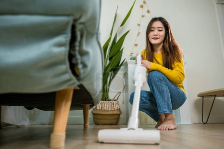 Téléchargez les photos : Housewife female dust cleaning floor under sofa or couch furniture with vacuum cleaner, Happy Asian young woman with accumulator vacuum cleaner at home in living room, household and housework concept - en image libre de droit