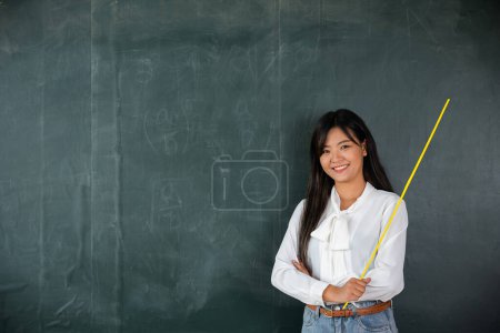 Foto de De vuelta al concepto escolar. Asiática profesora sonriendo con palo de madera en la pizarra delantera en la escuela en el aula, Feliz hermosa joven de pie mantenga puntero mirando a la cámara, Educación - Imagen libre de derechos