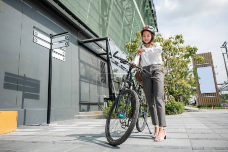Téléchargez les photos : Asiatique jeune femme d'affaires debout avec vélo vérifier l'heure sur la rue de la ville le matin avant d'aller au travail au bureau, portrait souriant femme vérifier montre, Trajet vers le travail, mode de vie motard - en image libre de droit