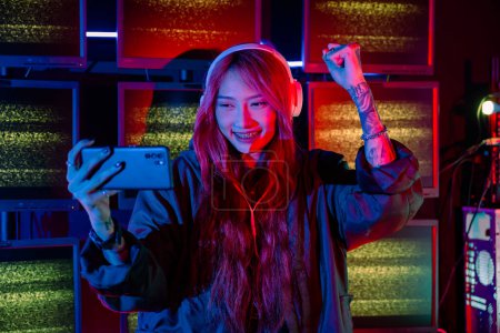Photo for Winning. Confused Asian woman wearing gaming headphones feeling excited at gaming room, Happy Gamer playing video game online with smart mobile phone with neon lights raises hands to wins celebrating - Royalty Free Image