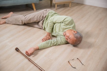 Téléchargez les photos : Homme âgé maux de tête couché sur le sol après être tombé il douleur et mal de l'ostéoporose, Homme âgé tombant sur le sol seul avec bâton de marche à la maison, Soins de santé et médicaments - en image libre de droit