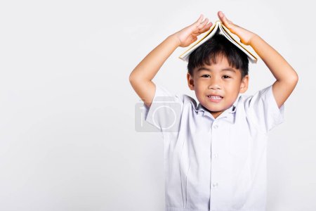 Photo for Asian adorable toddler smiling happy wearing student thai uniform red pants stand holding book over head like roof in studio shot isolated on white background, Portrait little children boy preschool - Royalty Free Image