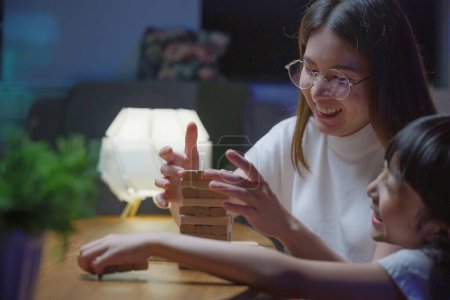 Photo for Smiling woman help teach child play build constructor of wooden blocks, Asian young mother playing game in wood block with her little daughter in home living room at night time before going to bed - Royalty Free Image