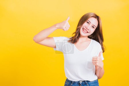 Photo for Portrait Asian Thai beautiful happy young woman smiling wear white t-shirt standing successful woman giving two thumbs up gesture sign in studio shot, isolated on yellow background with copy space - Royalty Free Image