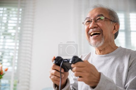 Foto de Asiático viejo hombre disfrutando de la celebración de joystick jugando videojuego en casa en la sala de estar, hombre maduro manos usando controlador de juego, Divertido en la jubilación anciano sonrisa sentado en el sofá juego de la vida - Imagen libre de derechos