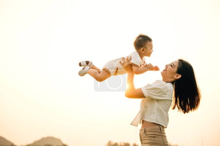 Photo for In a playful childhood moment, a happy Asian mom throws her little boy up into the sunny sky. The cheerful child enjoys the freedom of flying, while the joyful mother looks on with happiness and love - Royalty Free Image