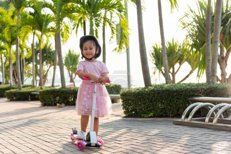 Téléchargez les photos : Scooter enfant. Joyeux Asiatique petite fille enfant porter un casque sûr en jouant rose kick board sur la route dans le parc à l'extérieur le jour de l'été, Jeux actifs pour enfants à l'extérieur, Sport pour enfants concept de mode de vie sain - en image libre de droit