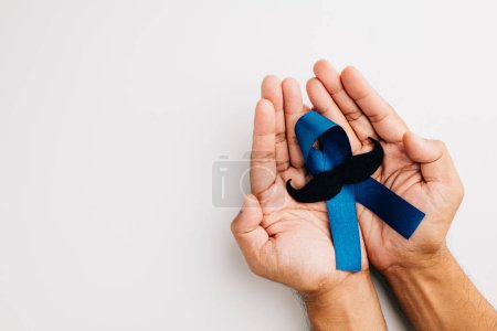 In November, mens health and Prostate cancer awareness take center stage. Mans hands embrace a light blue ribbon with a mustache on a blue background a symbol of support.