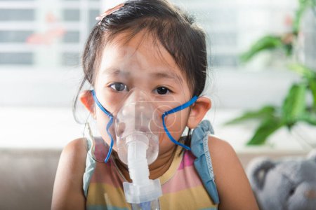 Photo for Kid girl making makes inhalation nebulizer steam sick cough at home, Asian Child using nebulizer mask equipment alone have smoke, stuffy nose and runny, oxygen spray inhaler therapy, Health medical - Royalty Free Image
