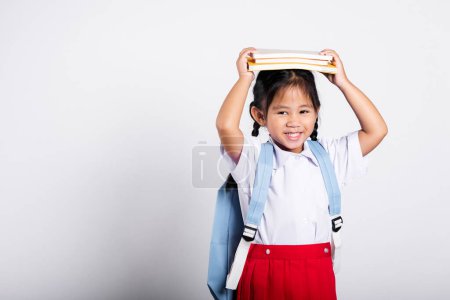 Foto de Asiático adorable niño sonriendo feliz desgaste estudiante tailandés uniforme falda roja pie sosteniendo libro sobre la cabeza y gritando en estudio tiro aislado sobre fondo blanco, Retrato niña preescolar - Imagen libre de derechos