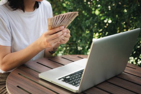 Téléchargez les photos : Gros plan mains de femme tenant beaucoup d'argent thai baht après avoir travaillé sur ordinateur portable à l'extérieur dans un café café, concept de carrière de réussite - en image libre de droit