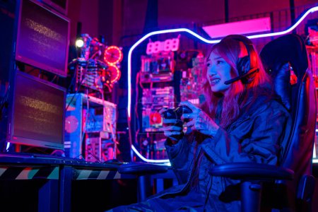 Téléchargez les photos : Jeune femme porter des écouteurs de jeu jouant salle de jeux console de jeux esports, Gamer en utilisant le contrôleur joystick pour tournoi virtuel joue jeu vidéo en ligne avec des néons d'ordinateur - en image libre de droit