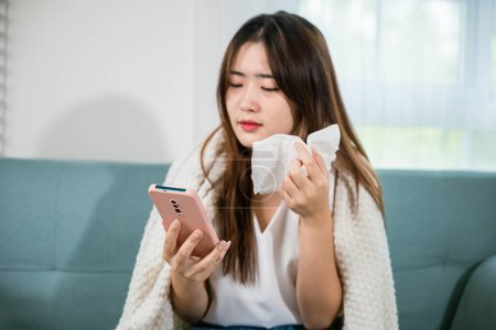 Foto de Asian young woman sick sitting down on sofa with cold blowing her nose and checking smart mobile phone to cell to doctor online, depressed sad sick female holding tissues and smartphone in living room - Imagen libre de derechos
