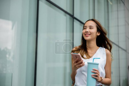 Foto de Concepto de estilo de vida urbano: Mujer caminando y usando su teléfono mientras sostiene una taza de vaso en una bulliciosa calle - Imagen libre de derechos