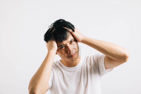 Foto de Un hombre asiático, abrumado por el estrés, sostiene su cabeza con dolor con una expresión triste. Estudio de tiro aislado en blanco, transmitiendo la sensación de tensión y enfermedad. - Imagen libre de derechos