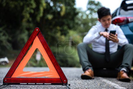 Photo for Efficiency and confidence on display as a young businessman changes the wheel of a broken car, while a worried mature man looks on. A red triangle sign warns drivers of the obstacle ahead. - Royalty Free Image