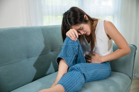 Photo for Asian young woman unhappy closing eyes with stomach ache on sofa in living room, female pain lying on couch at home, suffering from strong abdominal painful colon problem feeling unwell - Royalty Free Image