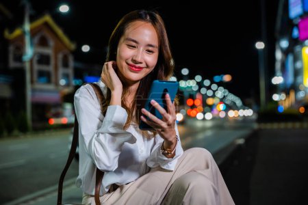 Foto de Mujer asiática en una calle de la ciudad por la noche combina a la perfección el trabajo y la vida personal mediante el uso de una computadora portátil y teléfono móvil mientras camina y habla. - Imagen libre de derechos