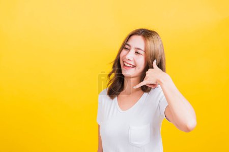 Photo for Portrait Asian Thai beautiful happy young woman wear white t-shirt standing smiling white teeth, making call me gesture by finger hand isolated on yellow background, with copy space - Royalty Free Image