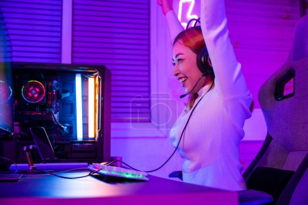 Foto de Ganar la victoria. Jugador asiático que juega el videojuego en línea excitado en la computadora de escritorio PC luces coloridas del neón led, mujer joven en auriculares del juego usando la computadora ella feliz acertado, concepto del E-Sport - Imagen libre de derechos