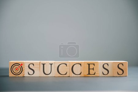 Symbolische Darstellung des Zielerfolgs mit einem Holzwürfel über dem Wort Erfolg auf einem Holztisch. Eingefangen auf einem minimalistischen grauen Hintergrund.