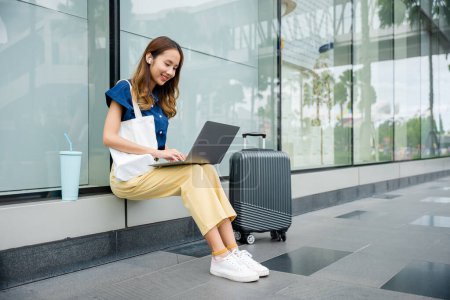 Photo for A modern woman works remotely using her laptop while sitting on a ledge at the airport, looking professional and productive. - Royalty Free Image
