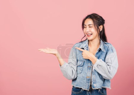 Téléchargez les photos : Portrait Asiatique belle jeune femme souriante usure denim debout pointant du doigt sur fond rose, Thai heureux visage excité point féminin dans vide regardant de côté avec de l'espace de copie pour le texte - en image libre de droit