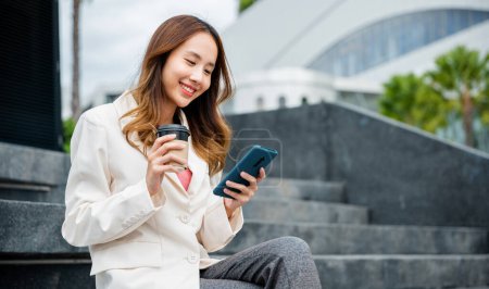 Téléchargez les photos : Lifestyle woman business people using mobile smart phones in the city outdoors office building. Young Asian female wearing blue shirt while use cellphone with holding coffee cup sitting on staircase - en image libre de droit