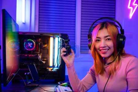 Foto de Smiling gamer using joystick controller for tournament plays online video game with computer neon light, woman wear gaming headphones playing live stream esports games console looking to camera - Imagen libre de derechos