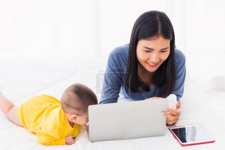 Téléchargez les photos : Asiatique nouvelle jeune belle mère trouver et lit des informations sur un ordinateur portable pour le mélange de lait sur une bouteille dans la chambre à coucher à la maison - en image libre de droit