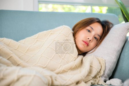 Foto de Asiática enferma mujer fría fiebre alta cubierta con manta descansando durmiendo en casa, mujer joven tiene problemas con dolor de cabeza acostado en el sofá en la sala de estar, chica enferma infeliz, persona cansada - Imagen libre de derechos