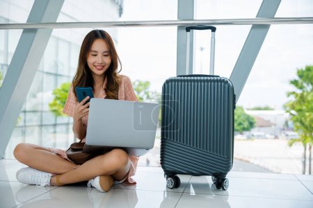 Foto de Una joven con un portátil y una maleta, trabajando en su teléfono en el aeropuerto. Es una productiva nómada digital, aprovechando al máximo su tiempo. La tecnología es su equipo. Concepto de viaje aéreo - Imagen libre de derechos