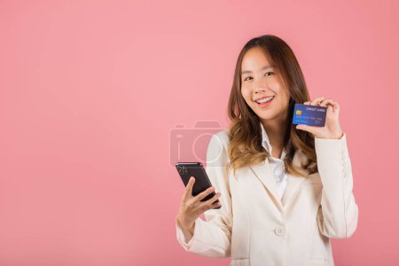 Téléchargez les photos : Heureux portrait asiatique belle jeune femme mignonne excitée souriant tenir téléphone mobile et carte bancaire de débit en plastique, studio tourné isolé sur fond rose, femme en utilisant smartphone achats en ligne - en image libre de droit