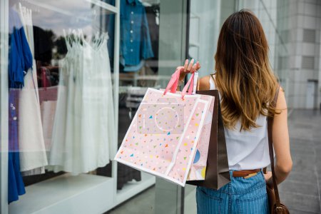 Photo for Young woman walking in front of clothing store with shopping bags, surrounded by busy urban buildings and bustling transportation. She is a trendy shopper, always following the latest fashion trends. - Royalty Free Image