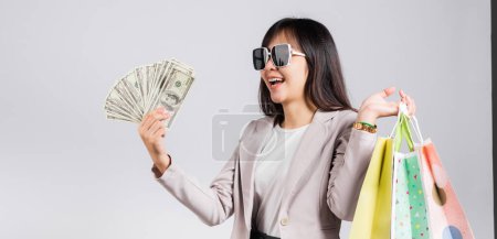 Téléchargez les photos : Femme avec des lunettes sourire client confiant tenant sacs à provisions en ligne multicolore et billets d'argent en dollars sur la main, heureux heureux asiatique jeune femme studio tourné isolé sur fond blanc - en image libre de droit