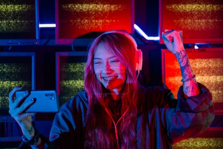 Photo for Winning. Young woman feeling excited at gaming room, Happy gamer people playing video game online with smart mobile phone with neon lights raising hands to wins celebrating, smile with smartphone - Royalty Free Image