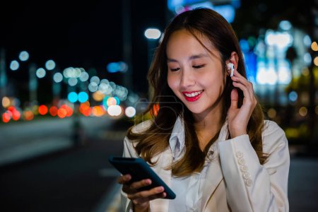 Foto de Un retrato de una joven hablando en su smartphone con auriculares, disfrutando de una conversación en una calle de la ciudad por la noche. El dispositivo portátil y la conexión inalámbrica lo hacen posible. Comunicación telefónica. - Imagen libre de derechos