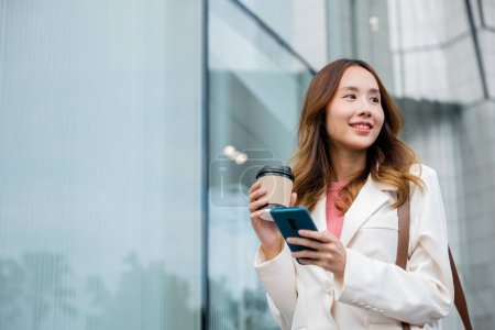 Téléchargez les photos : Jeune femme d'affaires asiatique indépendante regardant loin avec le sourire tout en utilisant un smartphone et tenant tasse de café sur la route à l'extérieur dans le parc urbain de la ville. Concept de travail à distance avec mode de vie flexible. - en image libre de droit