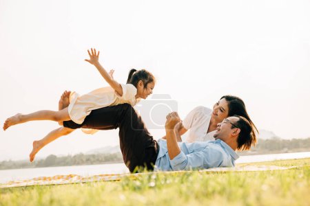 Foto de Una familia disfruta de momentos divertidos juntos. El padre sostiene a la hija en alto en el aire mientras se ríe y extiende sus brazos como alas, mientras que la madre observa con una sonrisa al aire libre. un precioso momento de diversión familiar - Imagen libre de derechos