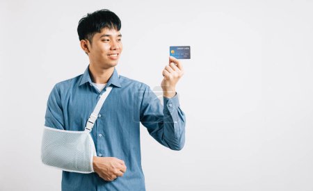 Photo for A man with broken arm displays confidence, donning support splint and using credit card for emergency medical bills. Happy Asian man in sling, isolated on white background, highlighting health care. - Royalty Free Image