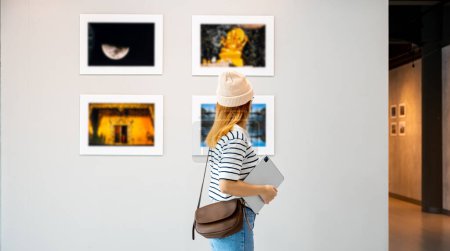Photo for Young person at photo frame hold digital book leaning against at show exhibit artwork gallery, Asian woman holding tablet at art gallery collection in front framed paintings looking pictures on wall - Royalty Free Image