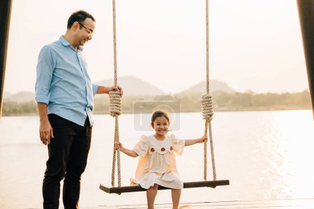 Photo for A happy Asian family spending quality time together on a sunny summer day at the playground, with the father pushing his cute daughter on a swing and both of them smiling and laughing with joy - Royalty Free Image